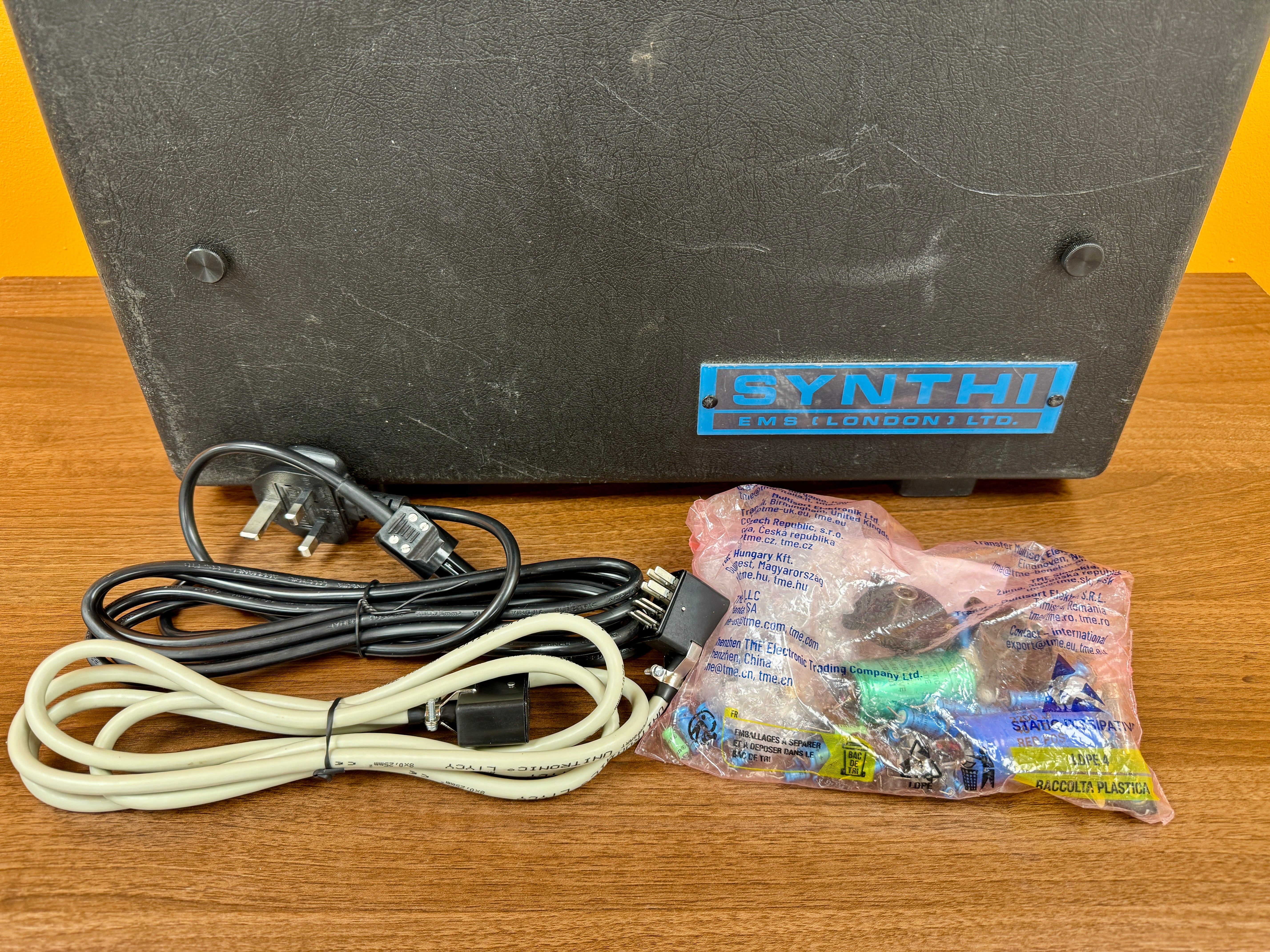 Black electronic device with cables and a small bag on a wooden surface, featuring the brand 'SYNTHI'.
