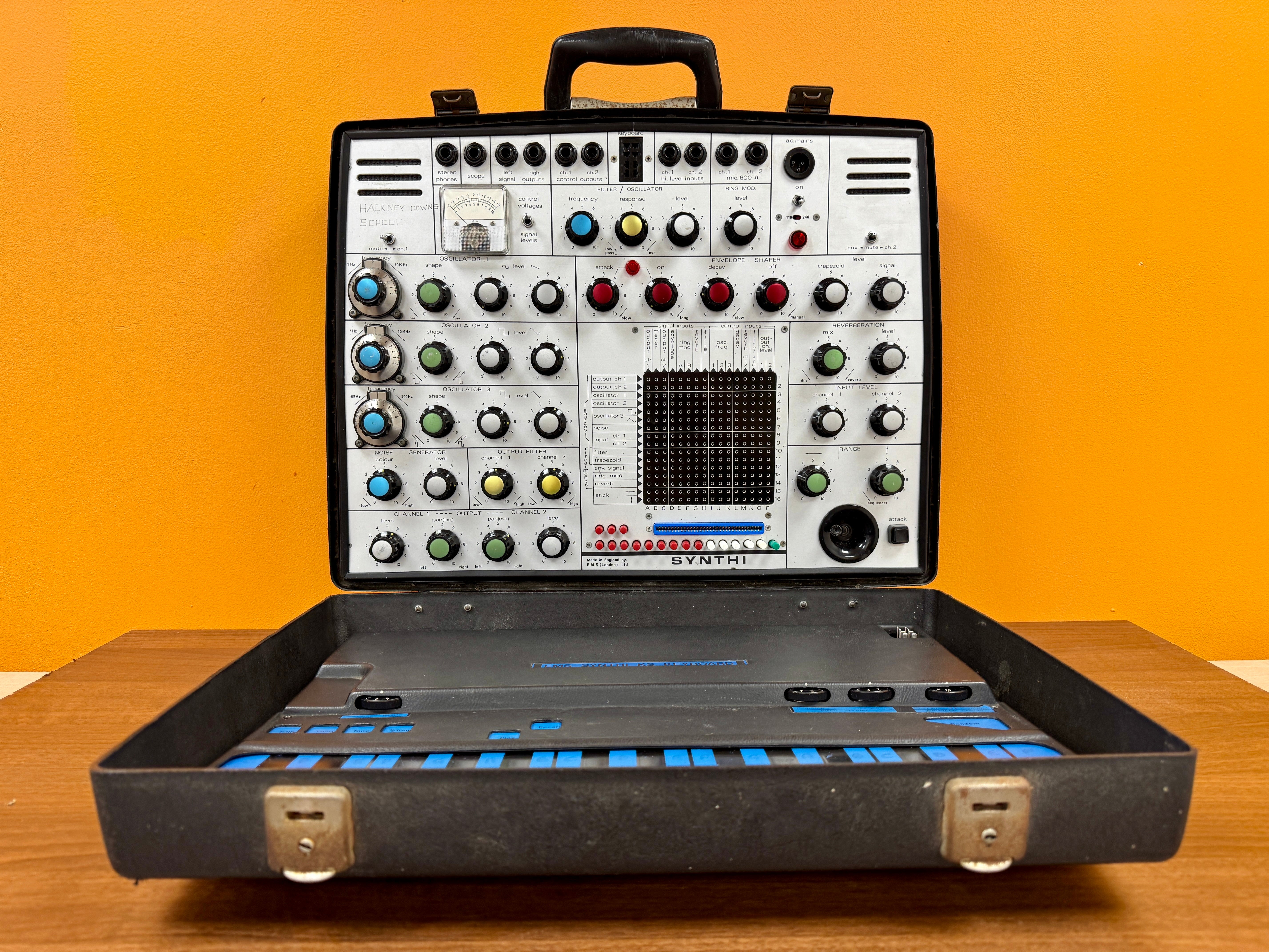 Portable electronic synthesizer with control panel and keyboard on a wooden surface against an orange background