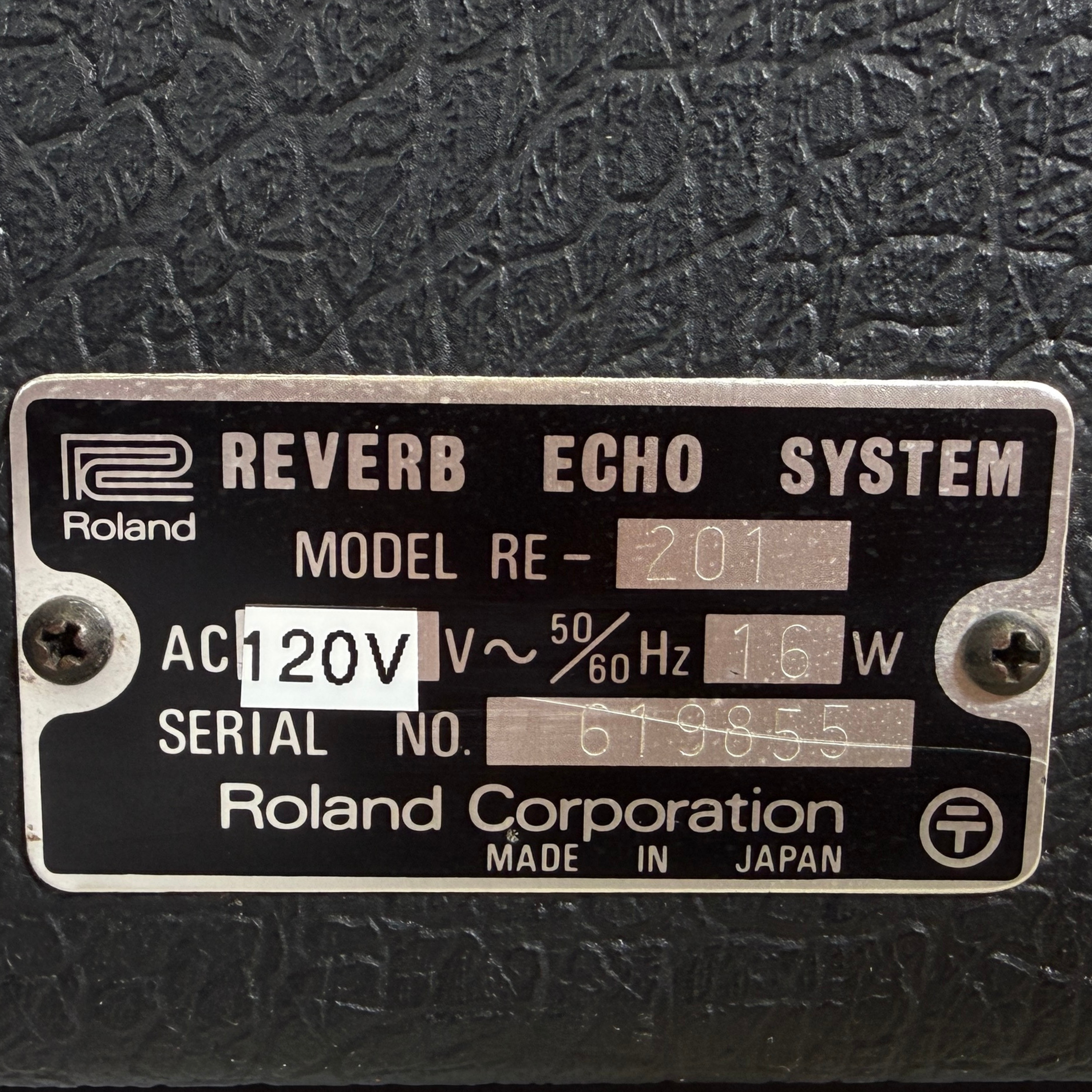 A close-up of a black metal product plate with silver text reading 'Roland Reverb Echo System' and 'Model RE-201', plus 'AC 120V' and serial number details, mounted on a dark textured surface.