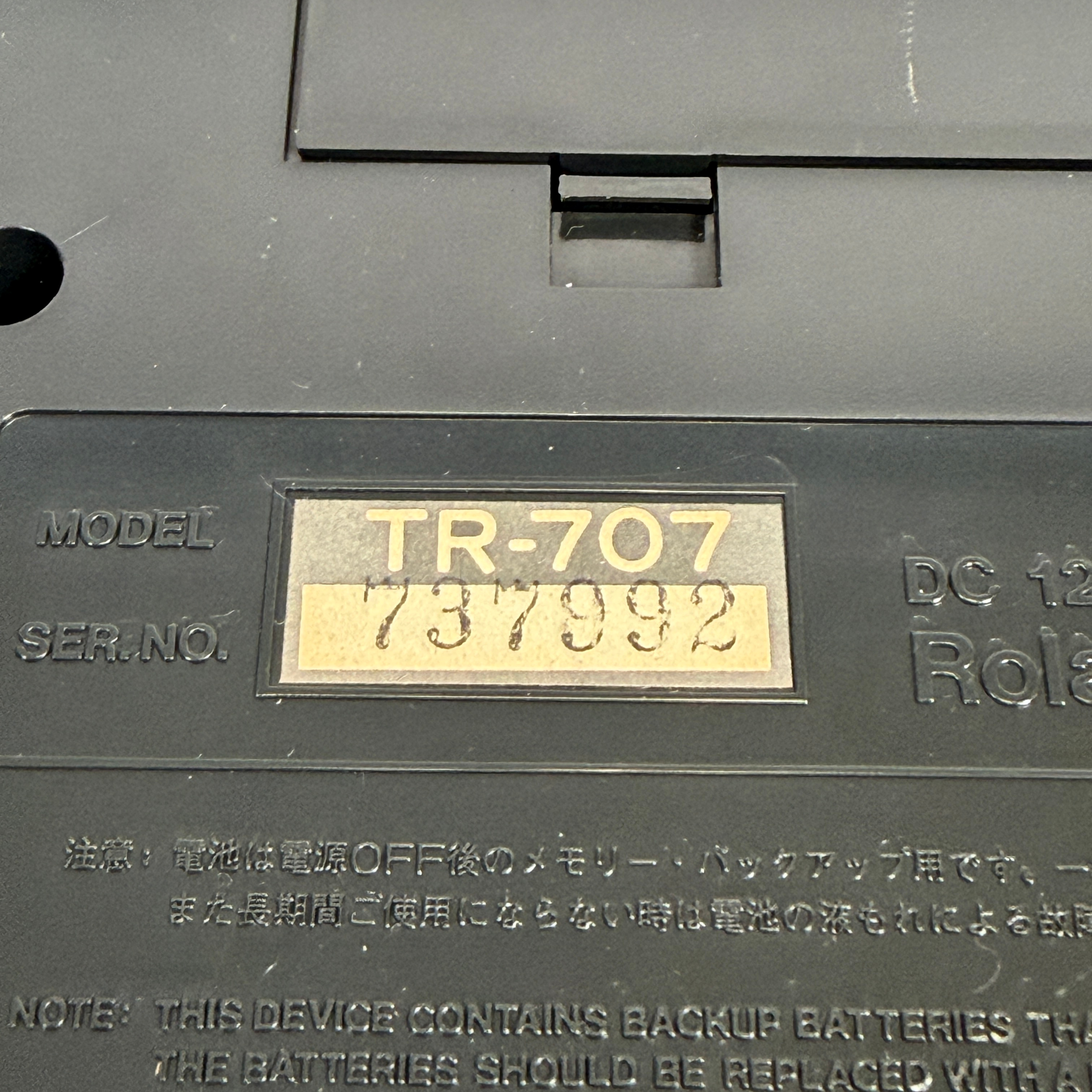 Close-up of a black plastic electronic device showing a yellowed serial number label with the text 'TR-707 737992' and embossed model and serial information.